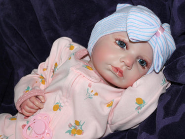 Lifelike reborn baby doll with blue eyes wearing a pink floral outfit and striped bow hat, realistic weighted newborn therapy doll